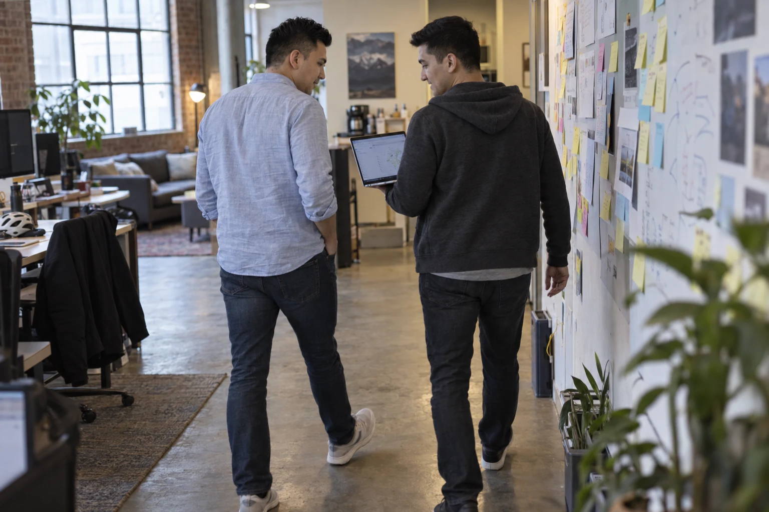 Illustrative workplace scene: Varis and Jasur discuss an open laptop while walking beside a wall of working notes.